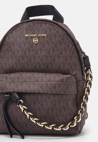 Zaino marrone con texture, caratterizzato da un prominente motivo a monogramma MK, logo dorato e manico con catena. Presenta un design arrotondato.