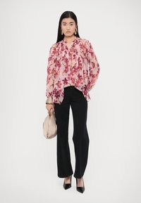Light pink floral blouse with long sleeves, flowing design, worn with black flared pants, holding a beige handbag, black pointed heels.