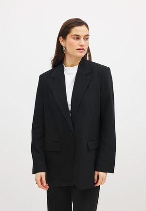 Blazer nero con una vestibilità sartoriale, chiusura a un bottone, rever a lancia e due tasche frontali. Indossato sopra una maglietta bianca.