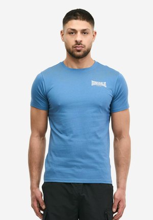 Uomo con capelli corti che indossa una maglietta aderente blu Lonsdale e pantaloni neri, in piedi contro uno sfondo chiaro e uniforme.