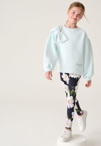 Lichtblauwe sweatshirt met een strikdetail op de schouder, gecombineerd met marineblauwe leggings met een bloemenpatroon, aangevuld met witte sneakers.