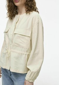 Svetlá béžová košeľa s gombíkmi a dvoma náprsníkovými vreckami. Má elastické manžety a šnúrku na stiahnutie v páse. Hladká, ľahká tkanina.