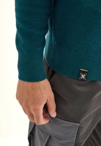 Pull en tricot teal avec un motif texturé, des poignets côtelés et une étiquette noire avec un accent jaune. Main reposant contre un pantalon cargo gris.