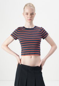 Crop top dengan garis horizontal merah, biru, dan navy, memiliki leher bulat, lengan pendek, dan desain yang pas, dipadukan dengan rok pleats hitam.