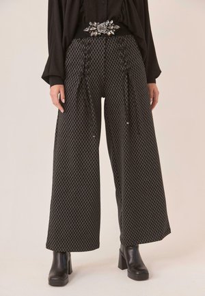 Pantalones anchos de pierna con estampado negro y detalles trenzados, combinados con botas negras de tacón y una blusa negra con hebilla de cinturón en forma de flor con joyas.