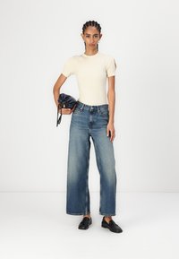 Μπεζ εφαρμοστό T-shirt, μπλε τζην παντελόνι με ψηλή μέση και φαρδιά πόδια, μαύρο loafers και μπλε τσάντα με κορδόνι. Μινιμαλιστικός, σύγχρονος σχεδιασμός.