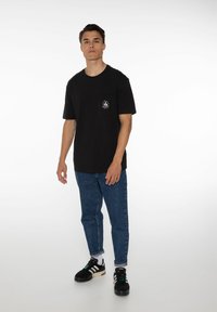 Zwart katoenen t-shirt met korte mouwen en een borstzakje met een wit logo. Gecombineerd met blauwe denimjeans en zwarte sneakers.