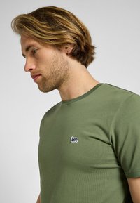 T-shirt z oliwkowej bawełny z okrągłym dekoltem i małym haftowanym logo "Lee" po lewej stronie klatki piersiowej. Gładki, dopasowany krój.