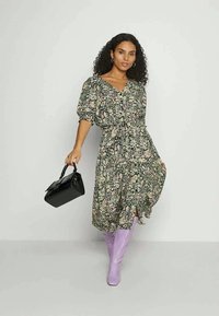 Mulher com cabelo encaracolado vestindo um vestido midi floral e botas lilás, segurando uma bolsa preta contra um fundo claro.