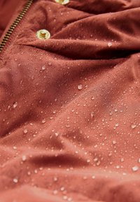 Wasserabweisender Stoff in Rostfarbe mit sichtbaren Tropfen. Hat eine glatte Textur und metallische goldene Hardware-Details.