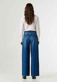 Wide-leg denim jeans in medium blue wash with a high waist, five pockets, and contrast stitching. Paired with a white long-sleeve shirt.