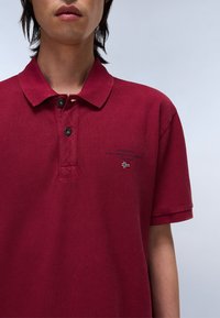 Burgundy színű, texturált pamut póló, két gombos gallérral, finom logó részlettel és zászló motívummal a mellkason.