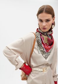 Beige satinblus med långa ärmar, kombinerad med en flerfärgad silkesscarf, röd läderhandske, brun väska och vit bälte med gulddetaljer.