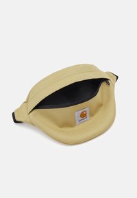 Riñonera beige de Carhartt con cremallera negra y parche del logo, parcialmente abierta, sobre fondo blanco.
