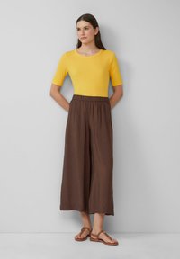 Femme se tenant debout avec les mains derrière le dos, portant un haut jaune à manches courtes, un pantalon large marron ample et des sandales marron, devant un mur gris.