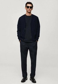 Marinblå cardigan, mörk t-shirt, skräddarsydda byxor och svarta skor. Snygga solglasögon. Materialen verkar vara släta med en strukturerad passform.