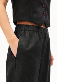 Person som har på sig mörkgrå byxor med elastisk midja, med händerna i fickorna, och en matchande kort väst med bruna knappar.