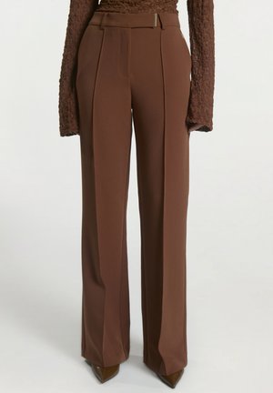 Bruine broek met wijde pijpen en plooien aan de voorkant, gedragen met een gestructureerde bruine top met lange mouwen en puntige bruine schoenen.