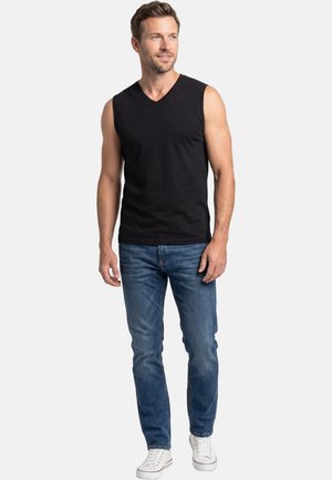 Man die staat en een zwart mouwloos V-halsshirt, blauwe spijkerbroek en witte sneakers draagt, licht zijwaarts kijkt en glimlacht.