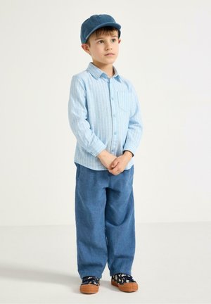Dreng iført blå kasket, lyseblå stribet skjorte, løse blå bukser og mønstrede sneakers, stående med hænderne foldet.