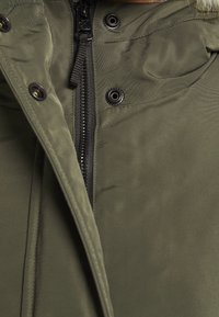 Kurtka oliwkowa z zamkiem błyskawicznym, zatrzaskami i miękką fakturą. Posiada kaptur i widoczne detale przeszyć.