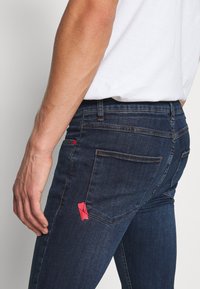 Los jeans de mezclilla azul oscuro presentan un ajuste slim, un diseño de cinco bolsillos y un detalle de costura roja en el bolsillo trasero. Tienen una textura suave y sin marcas de uso.