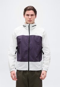 Νέος άνδρας που φοράει λευκό και σκούρο μοβ τζάκετ με φερμουάρ και ψηλό γιακά, μαζί με σκούρο παντελόνι, στέκεται μπροστά σε απλό λευκό φόντο.