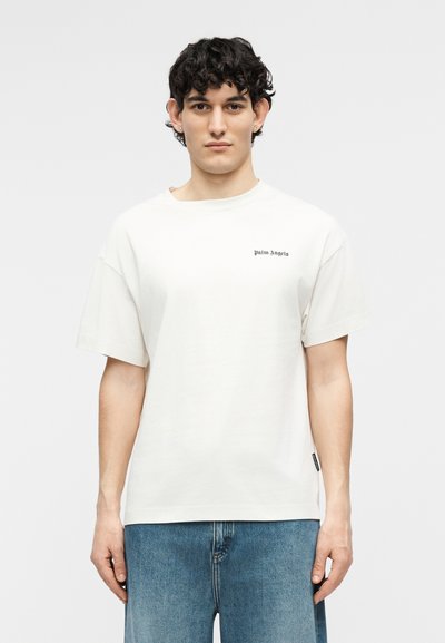 Νεαρός άνδρας με κοντά κατσαρά μαλλιά, φορώντας λευκό t-shirt Palm Angels και μπλε τζιν, στέκεται μπροστά σε απλό φωτεινό φόντο.