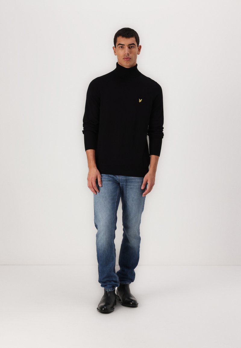 Un pull noir à col roulé avec des poignets côtelés, associé à un jean en denim bleu. Le mannequin porte des bottines noires et un petit logo jaune sur le pull.