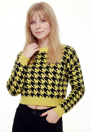 Ung blond kvinde, der blidt rører ved sin nakke, iført en gul og sort hundetandscrop sweater og sorte bukser på en hvid baggrund.