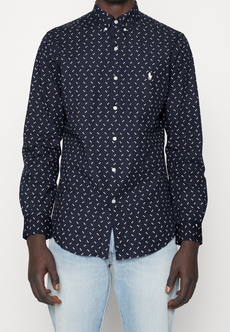 Homme portant une chemise boutonnée ajustée bleu marine avec de petits motifs à points blancs et un jean bleu clair, debout devant un fond uni.