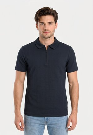 Uomo che indossa una polo blu scuro a maniche corte con cerniera frontale e jeans azzurri, in piedi davanti a uno sfondo semplice.