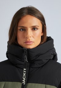 Doudoune noire et olive avec un col montant, fermeture éclair et détail de logo sur le devant. Texture lisse et design isolé.