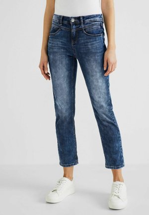 Femme portant un jean bleu court et des baskets blanches, debout devant un fond neutre.