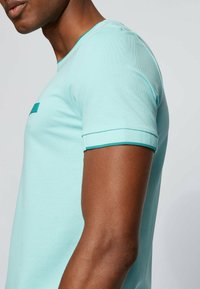 Světle tyrkysové tričko s krátkým rukávem, kulatým výstřihem, strukturovanou tkaninou, zeleným lemem na rukávech a malým logem na přední straně.