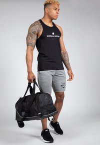 Fekete ujjatlan póló "GORILLA WEAR" logóval, szürke rövidnadrág és fekete sportcipő. Egy nagy fekete sporttáskát tart, amely hálós és szilárd anyagokból készült.
