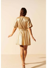 Robe métallisée dorée avec des manches courtes, taille cintrée et jupe évasée. Le tissu a une surface brillante et texturée, reflétant la lumière.