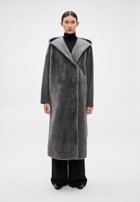 Dlhý sivý kabát s plyšovou textúrou, vybavený širokou kapucňou, zapínaním vpredu a svetlejším sivým podšívaním v kontrastnej farbe. Nosený cez čierne oblečenie.