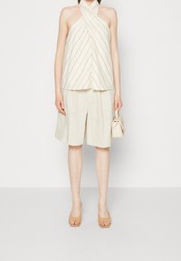 Top na zavazování za krkem ve světle krémové barvě s diagonálními béžovými pruhy, v kombinaci s béžovými lněnými šortkami. Modelka drží malou kabelku v neutrálních tónech.