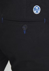 Czarne spodnie wykonane z wytrzymałej tkaniny, z niebieskim akcentem na guziku i przeszyciach. Zawierają naszywkę z napisem „North Sails” przy tylnej kieszeni.