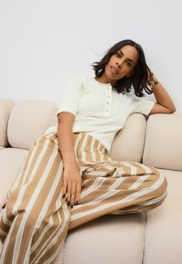 Light beige ribbed knit top with short sleeves and button placket, paired with wide-leg brown striped trousers, sitting on a cream sofa.