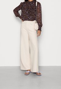Schwarze Bluse mit floralem Muster und langen Ärmeln, kombiniert mit beigefarbenen weit geschnittenen Hosen und lila Sandalen. Enthält eine kleine Umhängetasche.