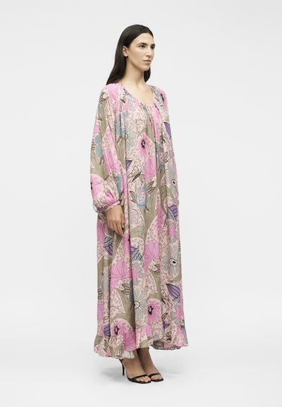 Γυναίκα που φοράει ένα μακρύ, φαρδύ ροζ φόρεμα με floral μοτίβο, μακριά μανίκια και βολάν στο τελείωμα, συνδυασμένο με μαύρα πέδιλα με ψηλά τακούνια, σε πλάγια στάση.