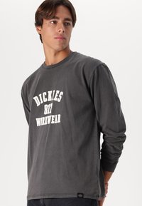 Jauns vīrietis valkā tumši pelēku garām piedurknēm kreklu ar uzrakstu "DICKIES 817 WORKWEAR", stāvot uz viendabīga balta fona.