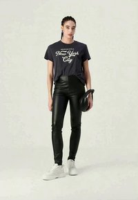 Γυναίκα που φοράει μαύρο μπλουζάκι "New York City", μαύρο δερμάτινο παντελόνι, λευκά αθλητικά παπούτσια και κρατάει μια μαύρη καμπύλη τσάντα.