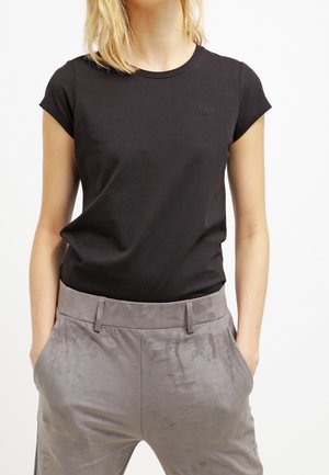 Vrouw draagt een nauwsluitend zwart T-shirt met korte mouwen, ingestopt in hoogtalgroene satijnen broek met handen in de zakken.