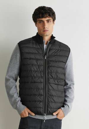 Mann trägt eine schwarze gesteppte Weste über einem hellgrauen Pullover, mit Händen in den Taschen, stehend vor einer schlichten hellen Wand.