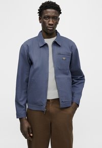 Νέος άνδρας που φοράει μπλε ζακέτα με φερμουάρ πάνω από λευκό πουκάμισο και καφέ παντελόνι, στέκεται με το ένα χέρι στην τσέπη μπροστά σε απλό φόντο.