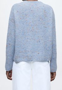 Personne portant un pull texturé bleu clair tacheté et un pantalon blanc, vue de dos sur un fond uni.