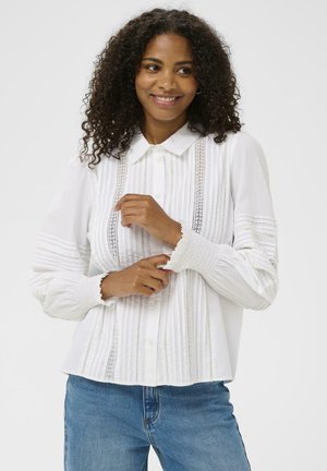 Femme souriante avec des cheveux bouclés portant une blouse blanche avec des détails en dentelle et des poignets smockés, associée à un jean bleu, sur fond uni.